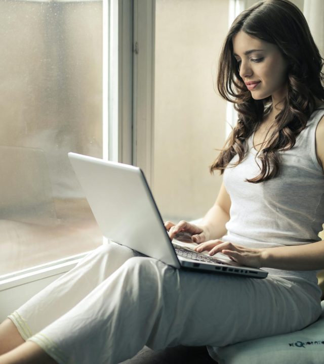 Adult woman sitting by window working remotely on a laptop indoors.